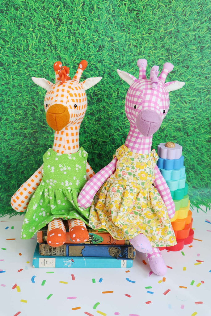 Two handmade giraffe dolls in dresses sitting on a stack of vintage books, made using the Noodle & Peach soft toy sewing pattern.