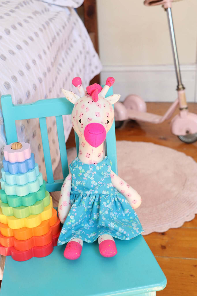 Handmade giraffe doll in blue floral dress seated on a turquoise chair, styled with pastel bedroom decor.