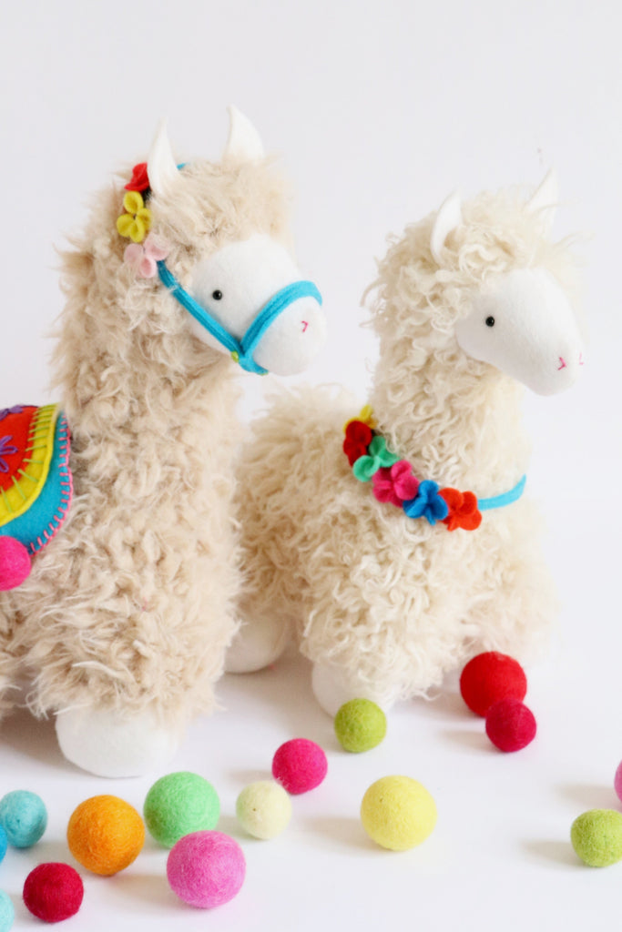 Handmade plush llama toys with curly fur, colorful felt saddle and garland, surrounded by bright felt balls. Soft toy sewing pattern example.