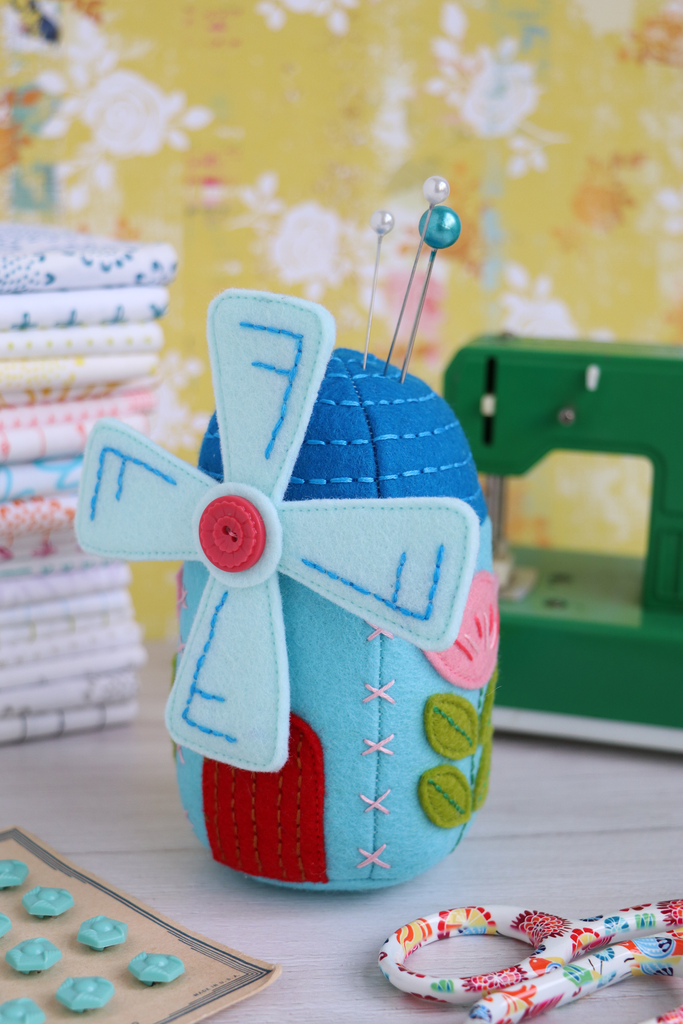 Blue windmill-shaped pincushion with a red button on a sewing-themed background.